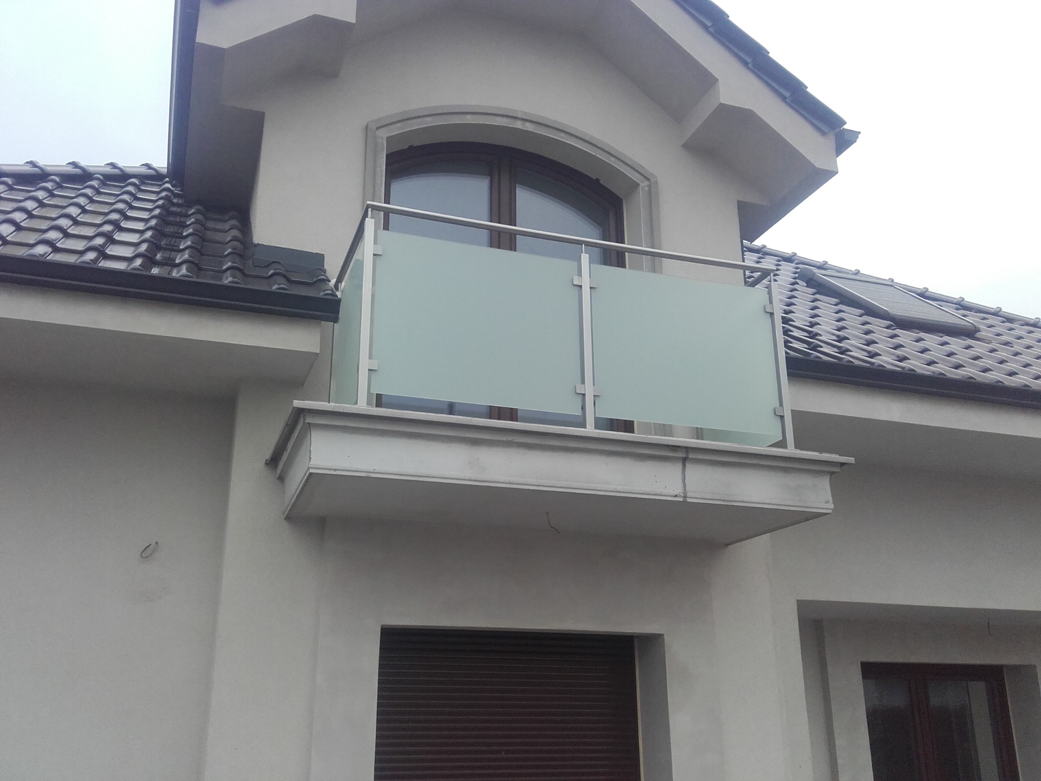 Balkon z przeszkloną balustradą ze stali nierdzewnej, widok z dołu, na tle szarej elewacji domu z brązowymi oknami i dachem z dachówki.