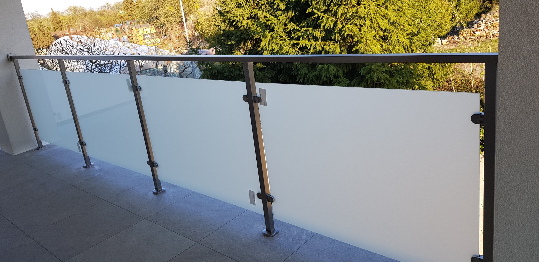 Nowoczesna balustrada balkonowa ze szkła matowego i metalu, widok z boku, na tle zieleni.