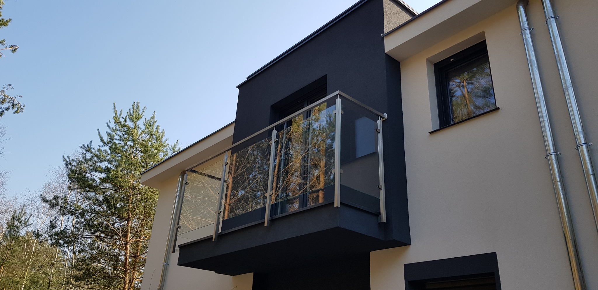 Nowoczesny balkon z balustradą szklaną i elementami ze stali nierdzewnej, widok z dołu na tle jasnego nieba i drzew