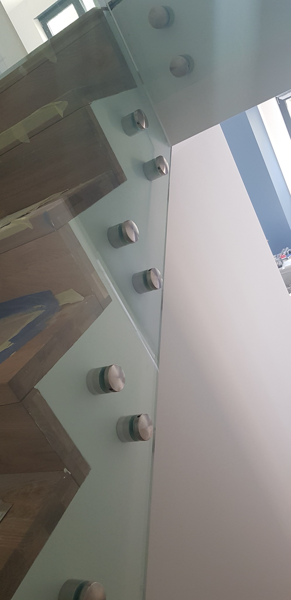 Szklana balustrada mocowana punktowo do drewnianych schodów, widoczne metalowe uchwyty i fragment białej ściany.