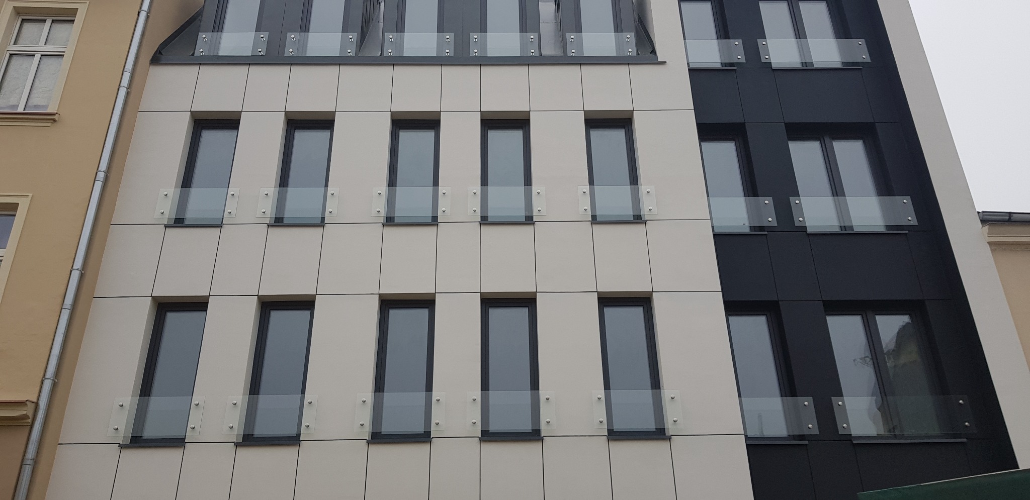 Elewacja budynku z oknami zabezpieczonymi szklanymi barierkami mocowanymi na metalowych wspornikach.