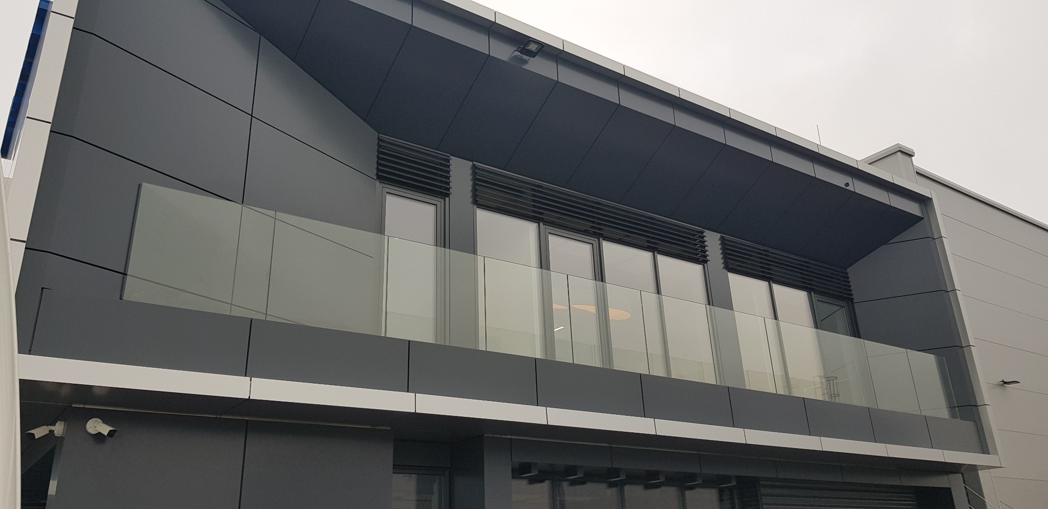Nowoczesny budynek biurowy z balkonem zabezpieczonym szklaną balustradą, widoczne kamery monitoringu na elewacji.