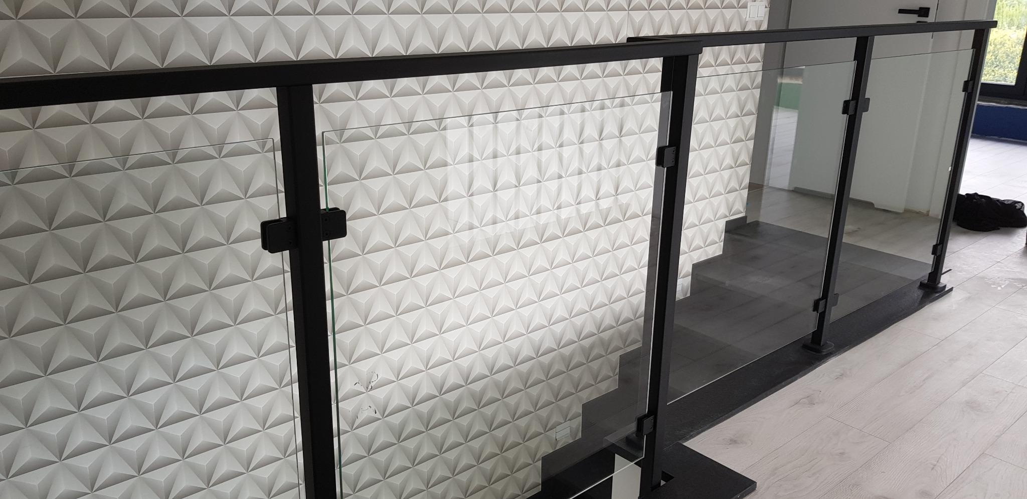 Szklana balustrada z czarnymi, metalowymi słupkami zamocowana na schodach, na tle ściany z trójwymiarową fakturą.