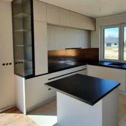 Nowoczesna kuchnia z wyspą i szklaną witryną, jasne fronty szafek, czarny blat, miedziana mozaika na ścianie, widok na dom przez okno, podłoga częściowo wykończona drewnem, częściowo płytkami.