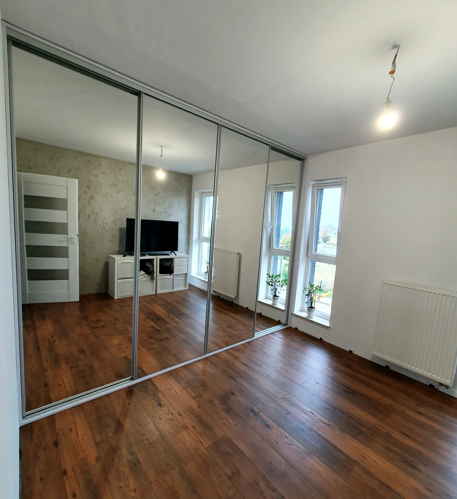 Duża szafa wnękowa z lustrzanymi drzwiami przesuwnymi w przestronnym pokoju z drewnianą podłogą i dwoma oknami, odbijająca wnętrze pokoju z telewizorem i białą komodą.