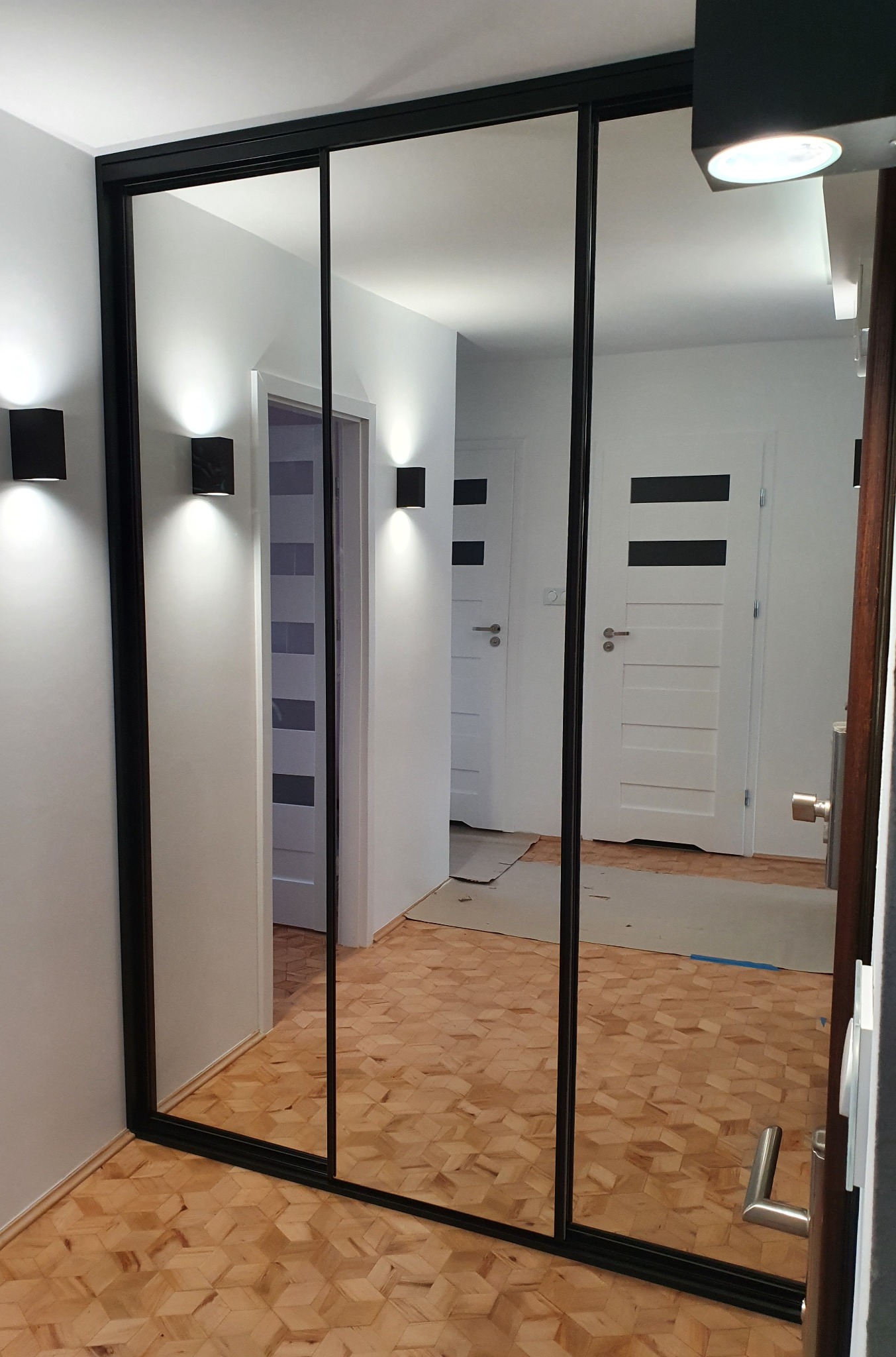 Trzydrzwiowa szafa wnękowa z lustrzanymi frontami i czarnymi obramowaniami, odbijająca wnętrze korytarza z parkietem w jodełkę i nowoczesnymi kinkietami.