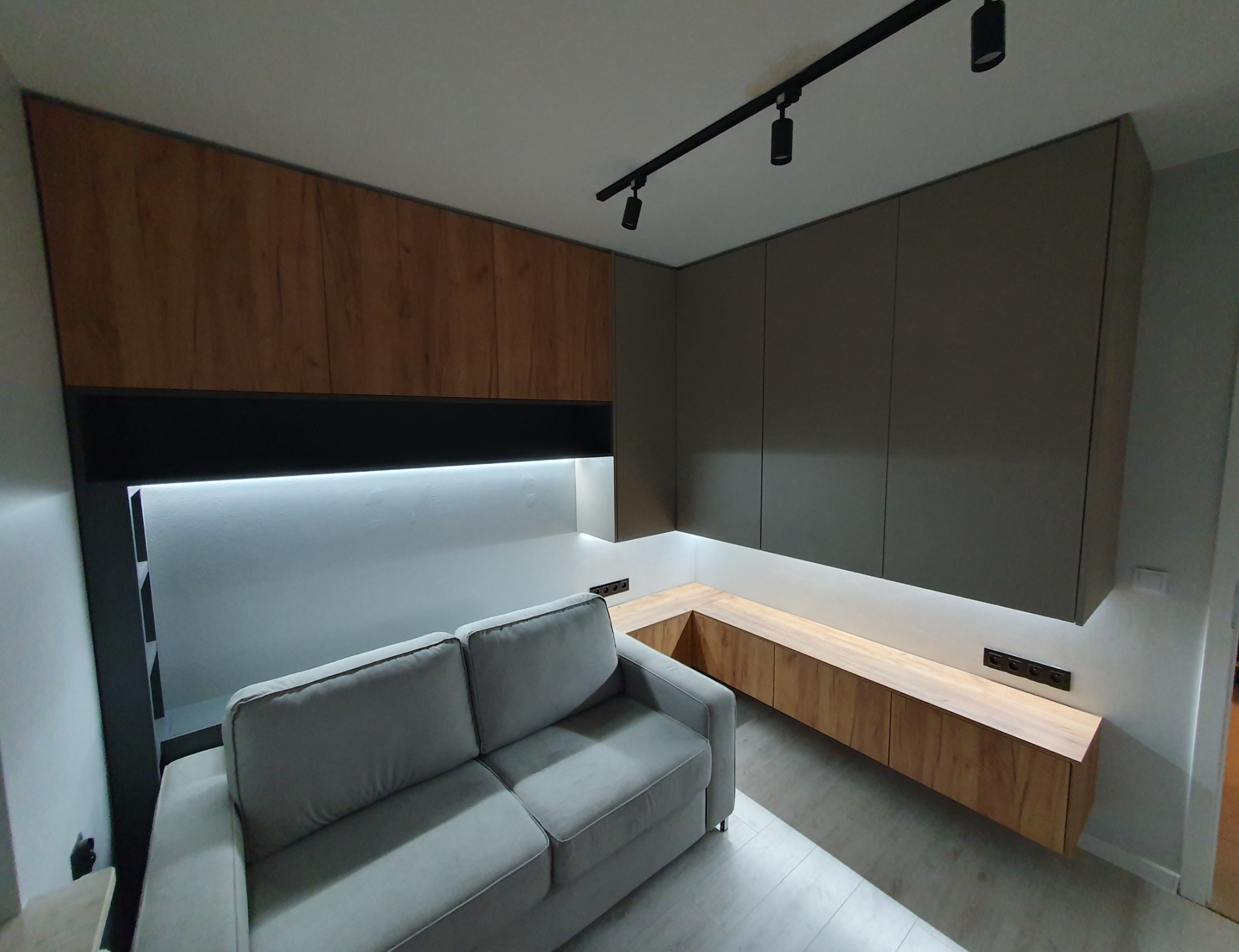 Nowoczesny salon z szarą sofą, zabudową meblową z oświetleniem LED i drewnianymi akcentami, widok na ścianę z gniazdkami.
