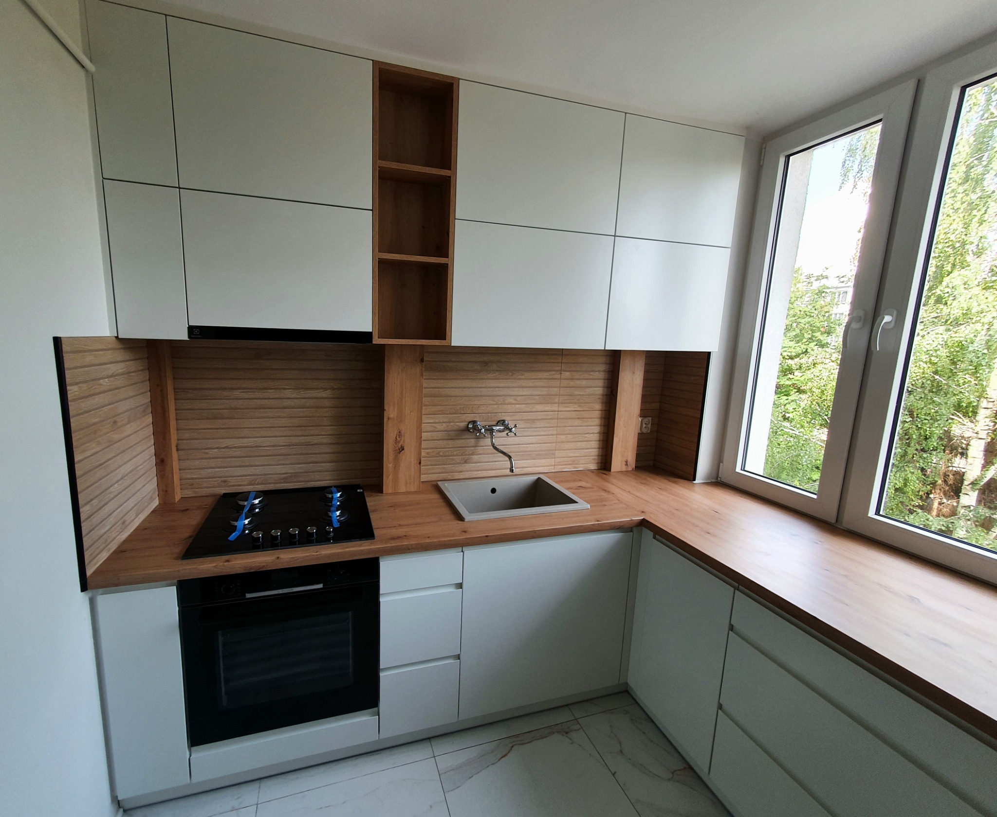 Nowoczesna kuchnia: białe szafki z drewnianymi akcentami, drewniany blat, płyta grzewcza z niebieską taśmą zabezpieczającą, szary zlew i duże okno z widokiem na zieleń.