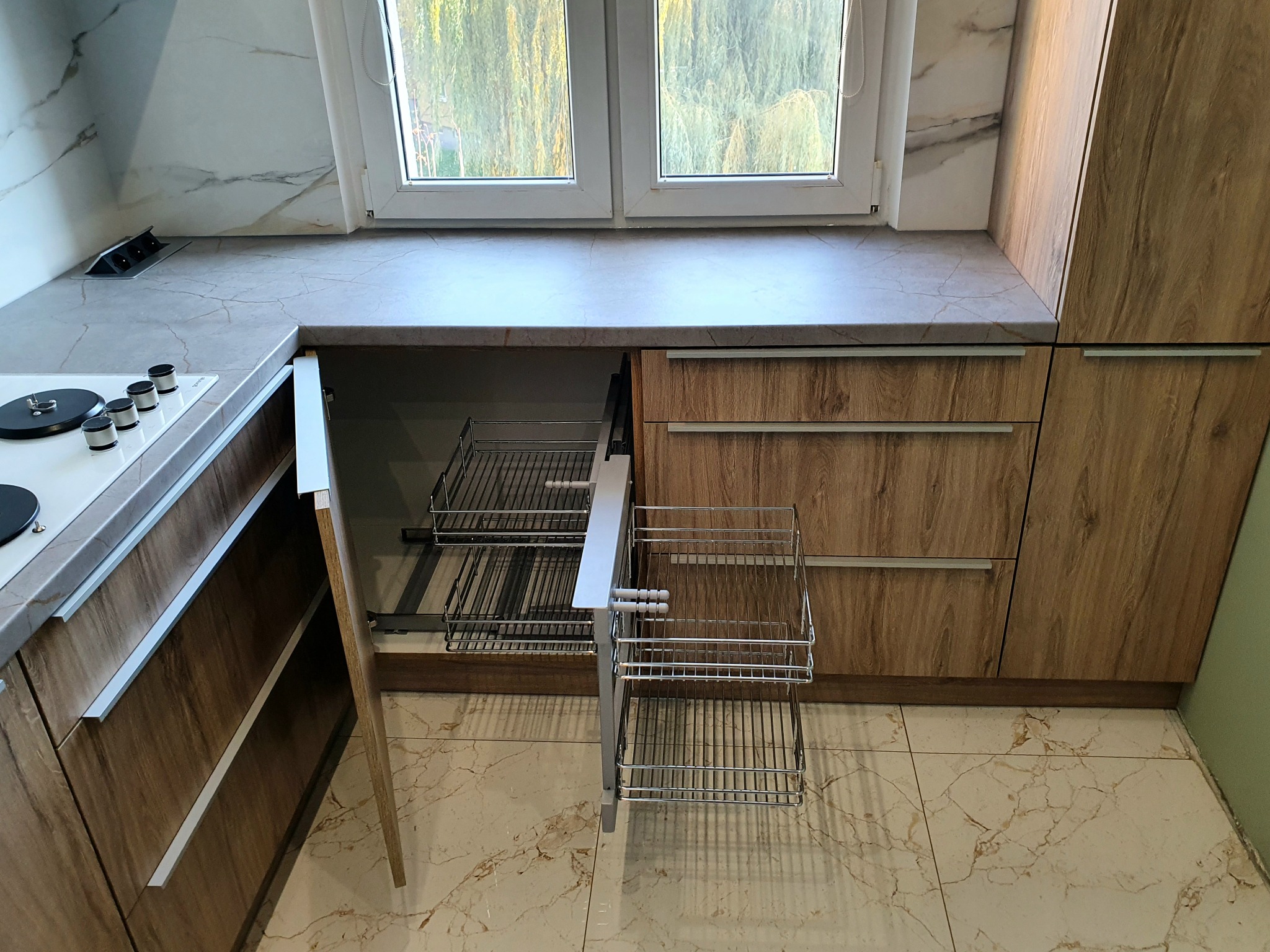 Wysuwany, metalowy kosz cargo w szafce z drewnopodobnymi frontami, część zabudowy kuchennej z blatem imitującym marmur, widok na okno i fragment płyty grzewczej.