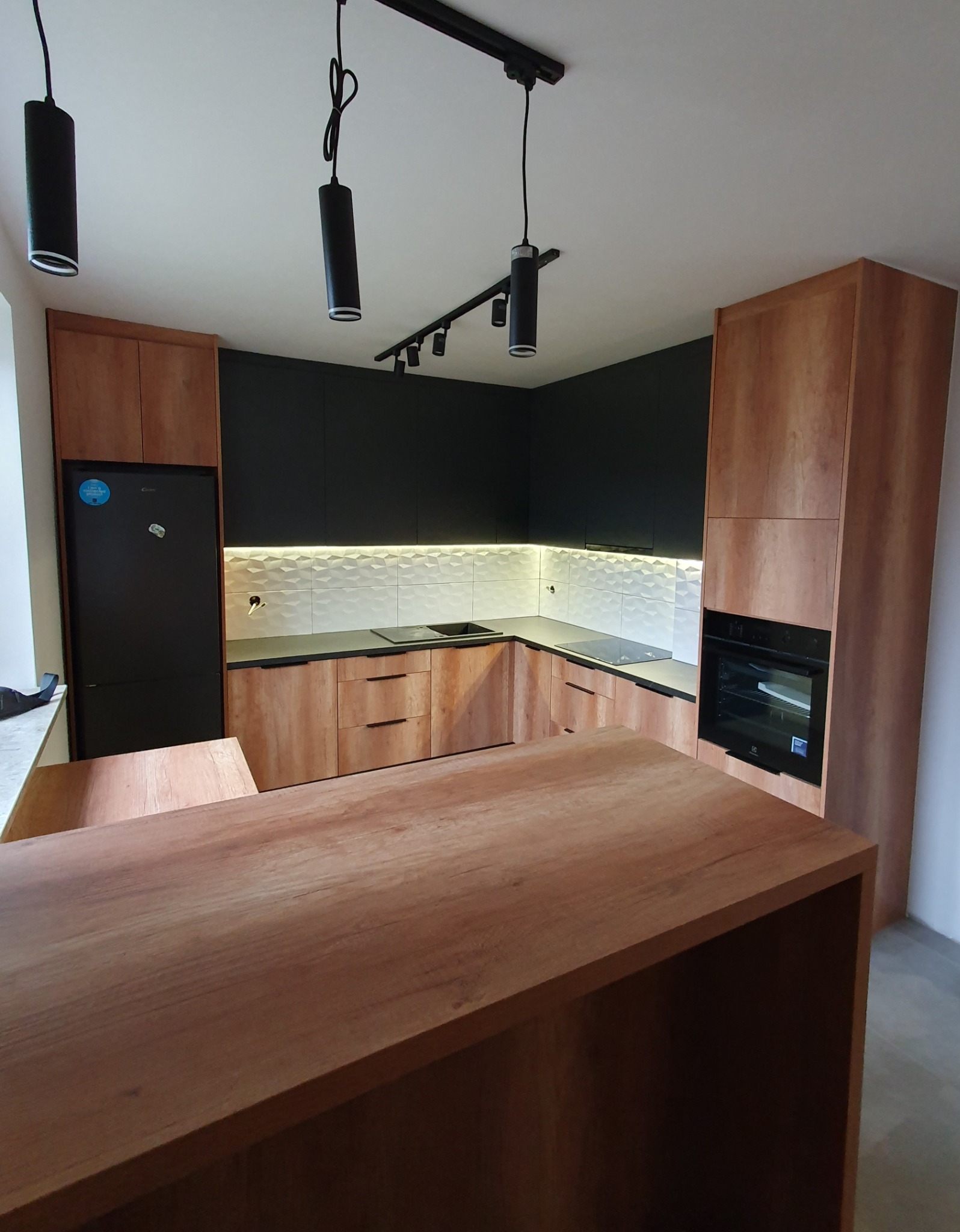 Nowoczesna kuchnia z drewnianymi frontami, czarnymi szafkami górnymi i wyspą kuchenną z blatem z litego drewna, oświetlona reflektorami sufitowymi.