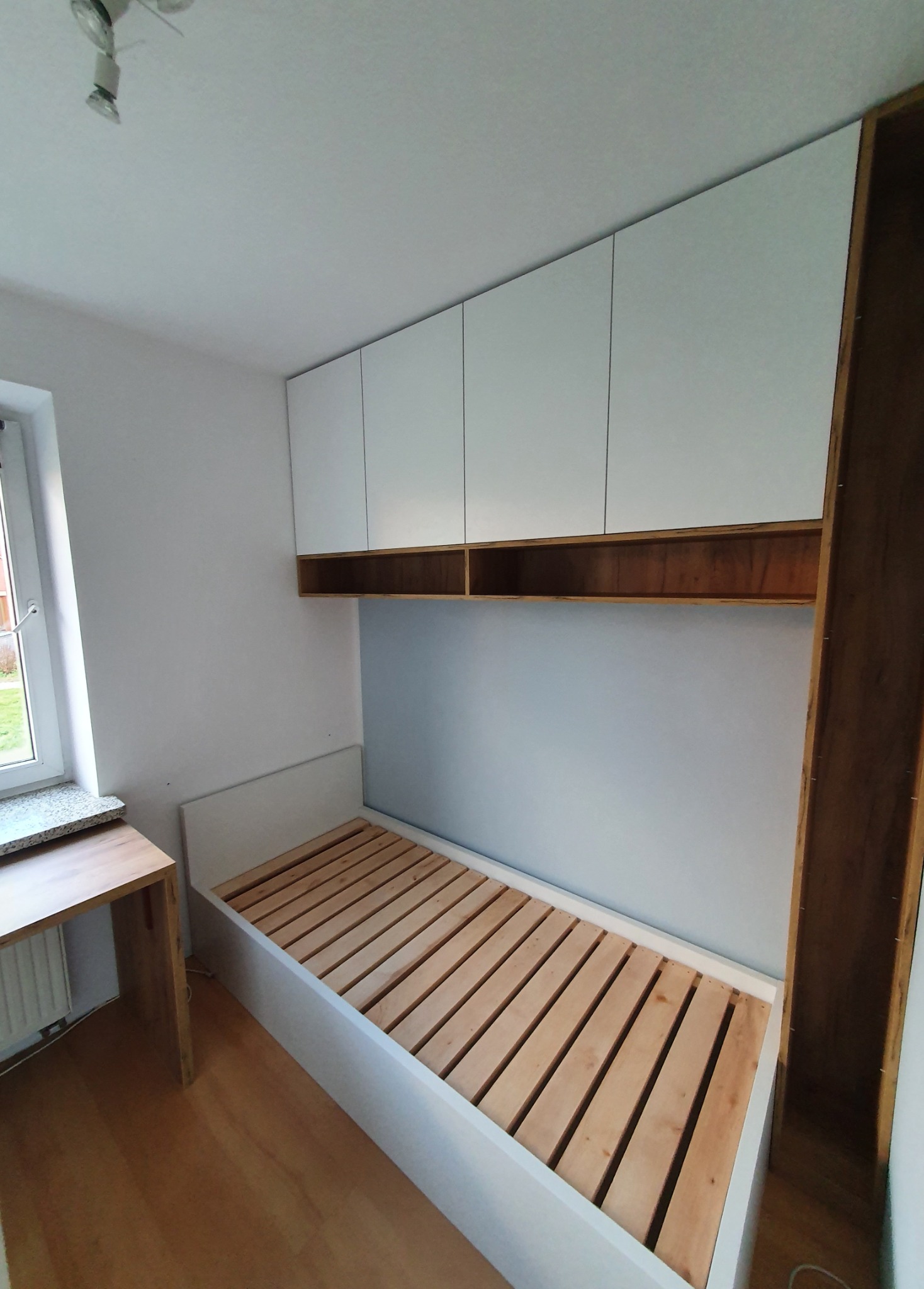 Pokój z łóżkiem bez materaca i szafkami wiszącymi nad nim, drewniany blat biurka przy oknie, widok na podłogę z paneli.