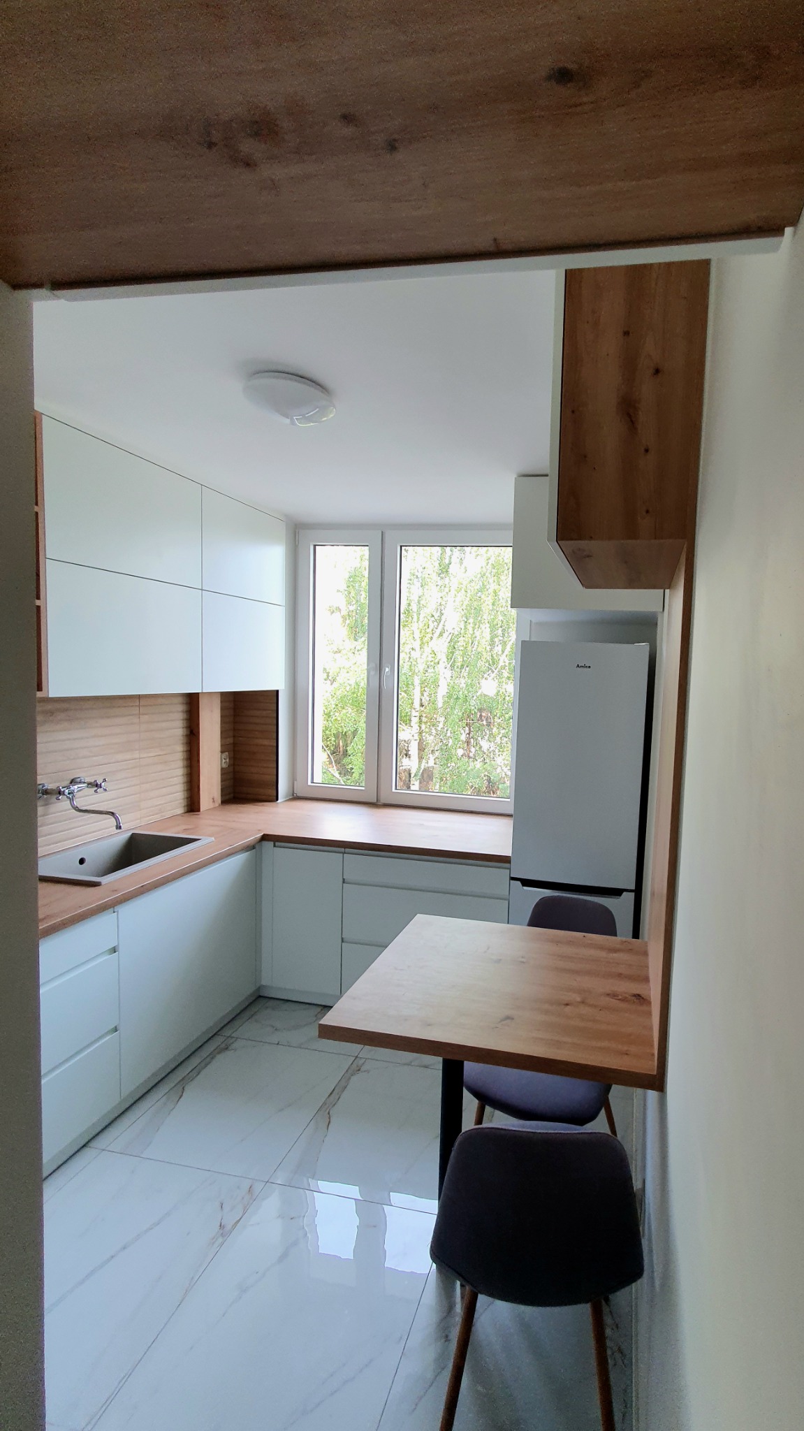 Jasna kuchnia z białymi frontami, drewnianym blatem i stołem z dwoma krzesłami, widok zza framugi drzwi.