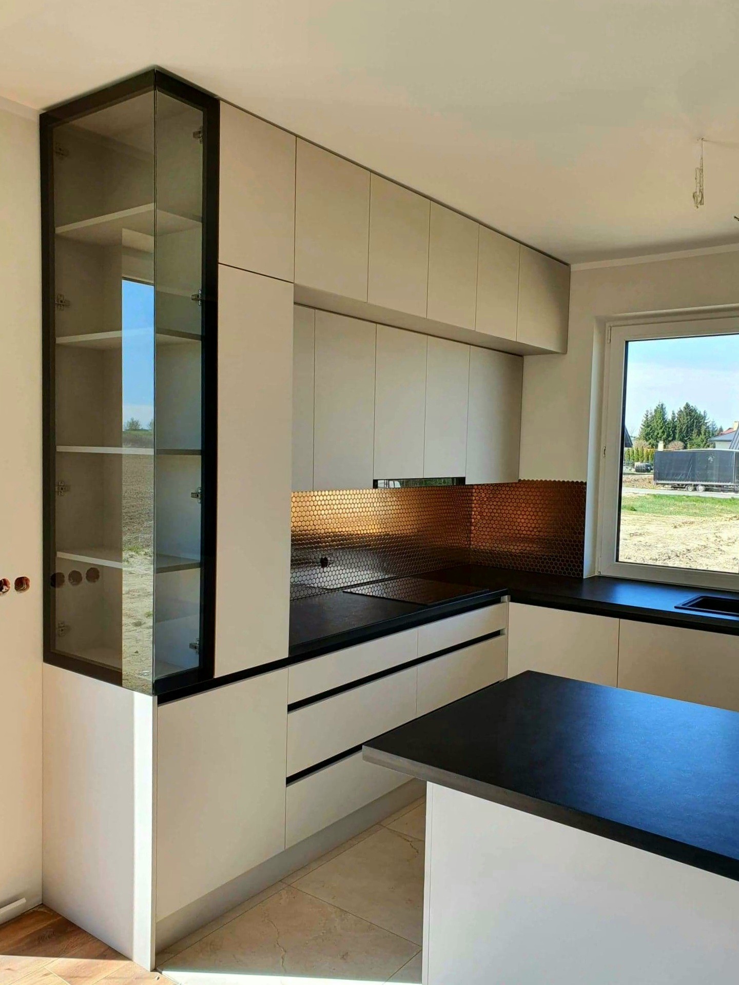 Jasna kuchnia z czarnym blatem, szklana witryna z czarną ramą, mozaika na ścianie i widok na budowę za oknem.