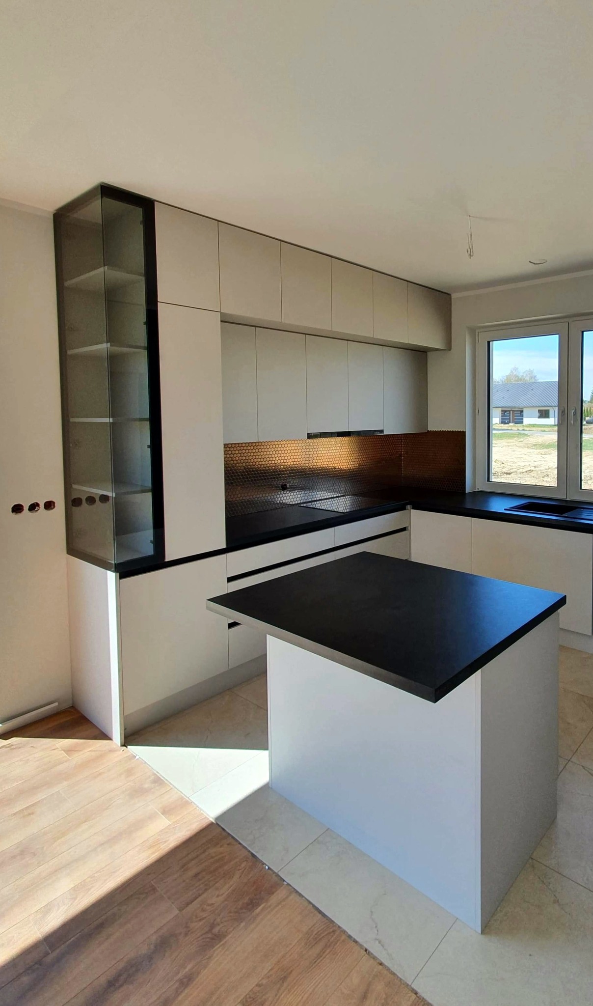 Nowoczesna kuchnia z wyspą i szklaną witryną, jasne fronty szafek, czarny blat, miedziana mozaika na ścianie, widok na dom przez okno, podłoga częściowo wykończona drewnem, częściowo płytkami.