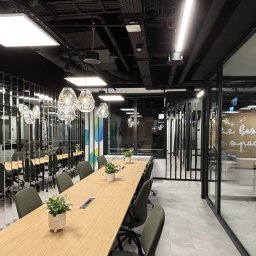 Apexim Bis Sp. z o.o. - Nowoczesna sala konferencyjna z długim stołem, krzesłami i wiszącymi lampami. Widoczne instalacje pod sufitem i szklane ściany działowe. Oświetlenie biurowe.