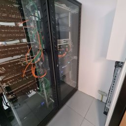 Apexim Bis Sp. z o.o. - Dwie szafy serwerowe z widocznymi kablami sieciowymi w kolorach pomarańczowym i zielonym, umieszczone w rogu pomieszczenia z szarą podłogą.