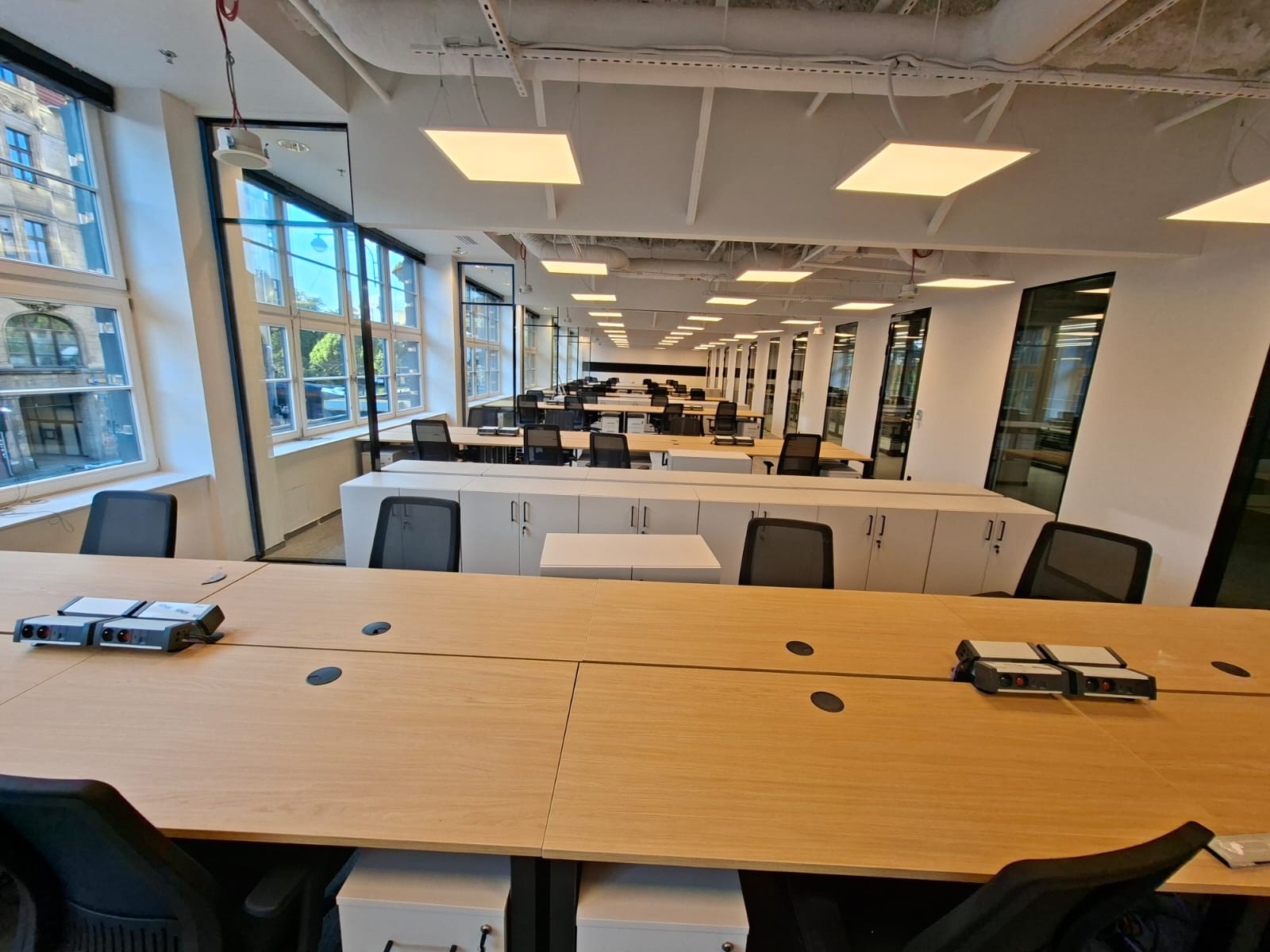 Nowoczesne biuro z rzędami biurek i kwadratowymi lampami sufitowymi. Widoczne okna z widokiem na budynek. Na biurkach zamontowane gniazda elektryczne.