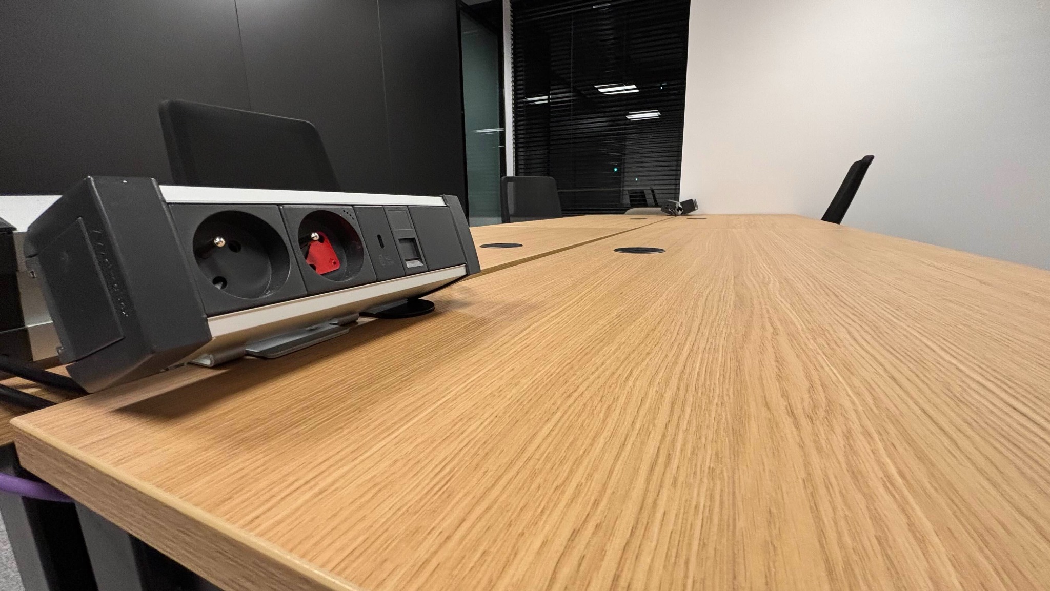 Wpuszczane gniazda elektryczne i porty USB w drewnianym blacie stołu konferencyjnego, widok z bliska, nowoczesne biuro z czarnymi i białymi ścianami w tle.