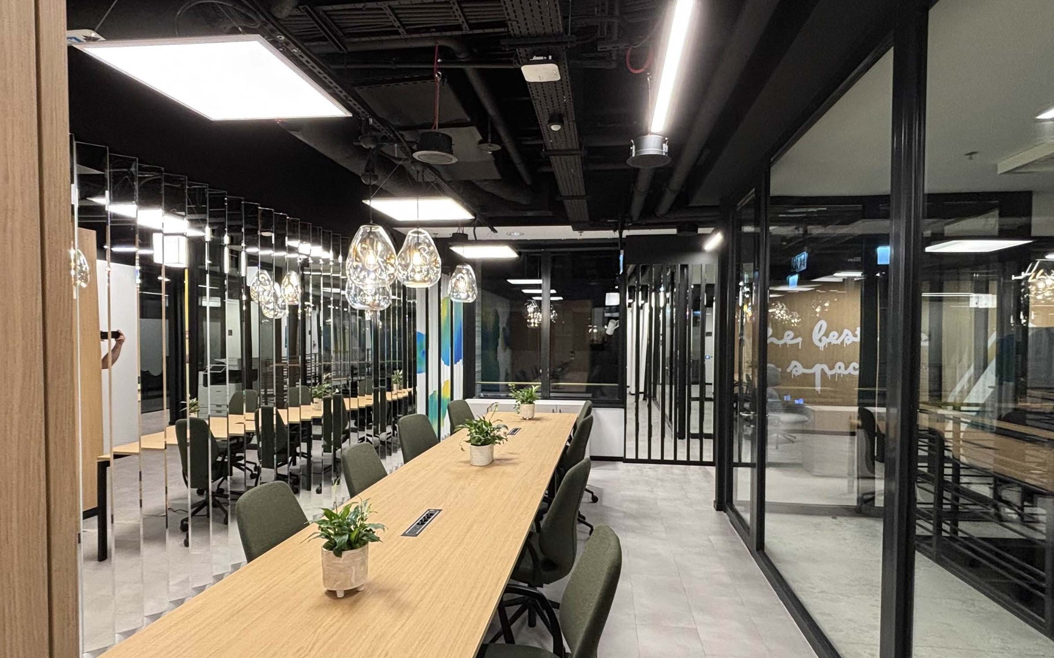 Nowoczesna sala konferencyjna z długim stołem, krzesłami i wiszącymi lampami. Widoczne instalacje pod sufitem i szklane ściany działowe. Oświetlenie biurowe.