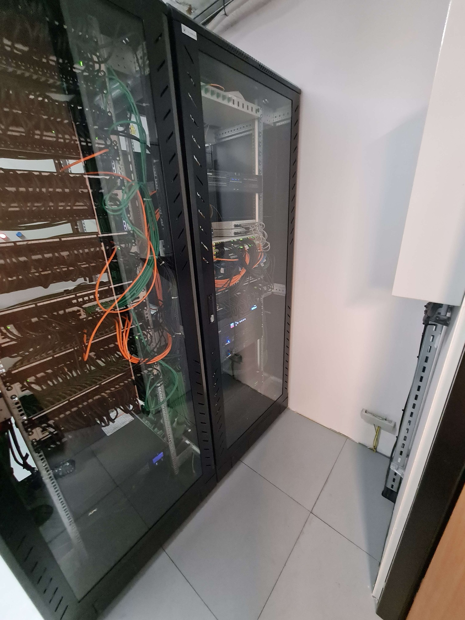 Dwie szafy serwerowe z widocznymi kablami sieciowymi w kolorach pomarańczowym i zielonym, umieszczone w rogu pomieszczenia z szarą podłogą.