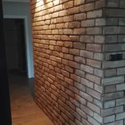 Ściana wykończona cegłą klinkierową z trzema czarnymi lampami ściennymi, oświetlającymi korytarz z drewnianą podłogą.