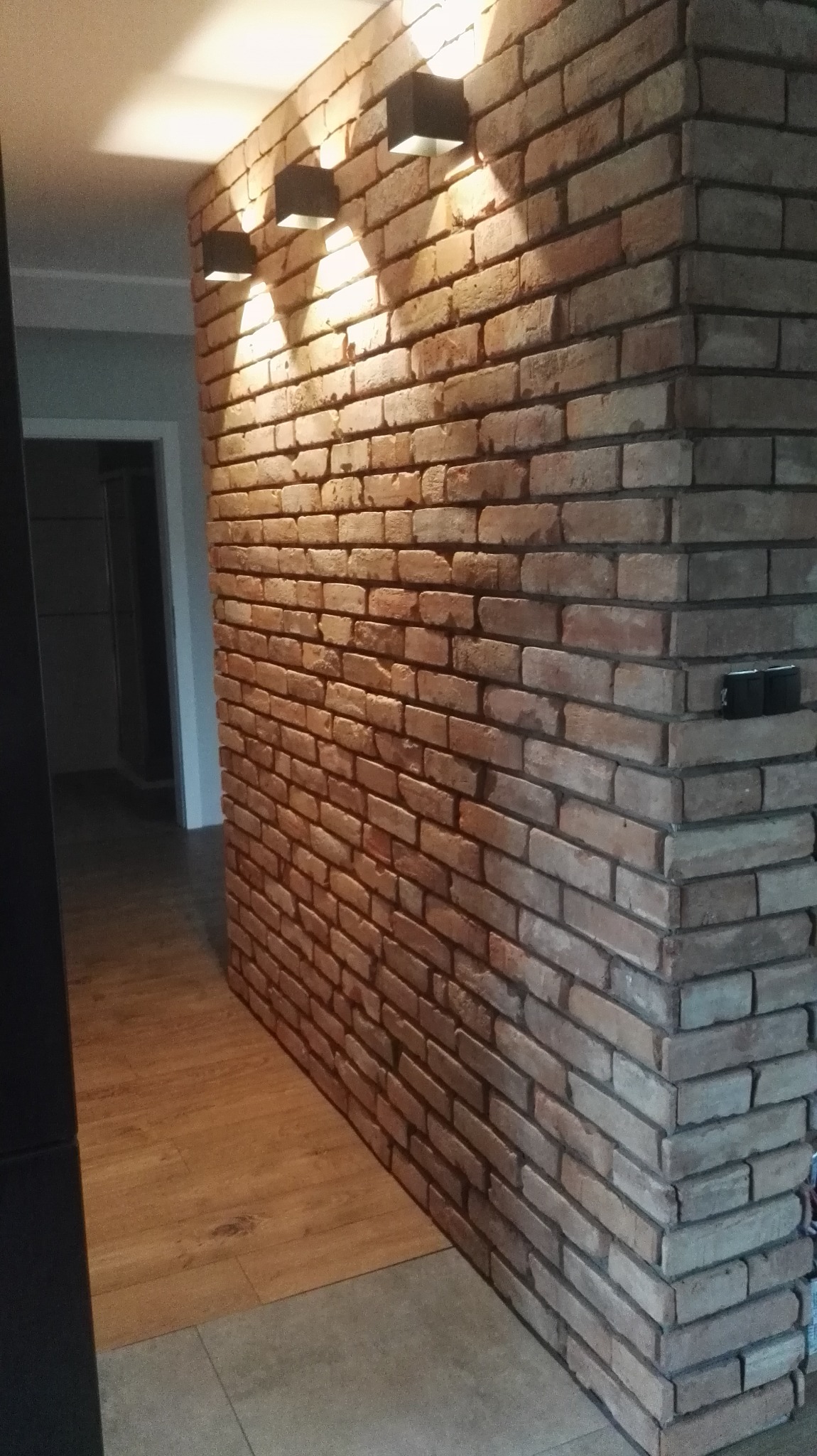 Ściana wykończona cegłą klinkierową z trzema czarnymi lampami ściennymi, oświetlającymi korytarz z drewnianą podłogą.