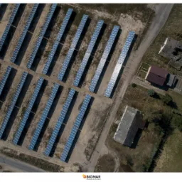 Widok z lotu ptaka na farmę paneli słonecznych z rzędami paneli ustawionymi równolegle, obok budynki w różnym stanie, w tym jeden zniszczony.