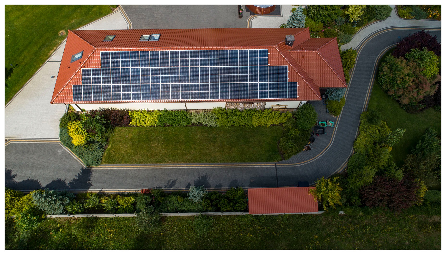 Widok z drona na dom z panelami słonecznymi na dachu, otoczony zadbanym ogrodem i brukowanym podjazdem, z widocznymi wózkami ogrodowymi obok domu.