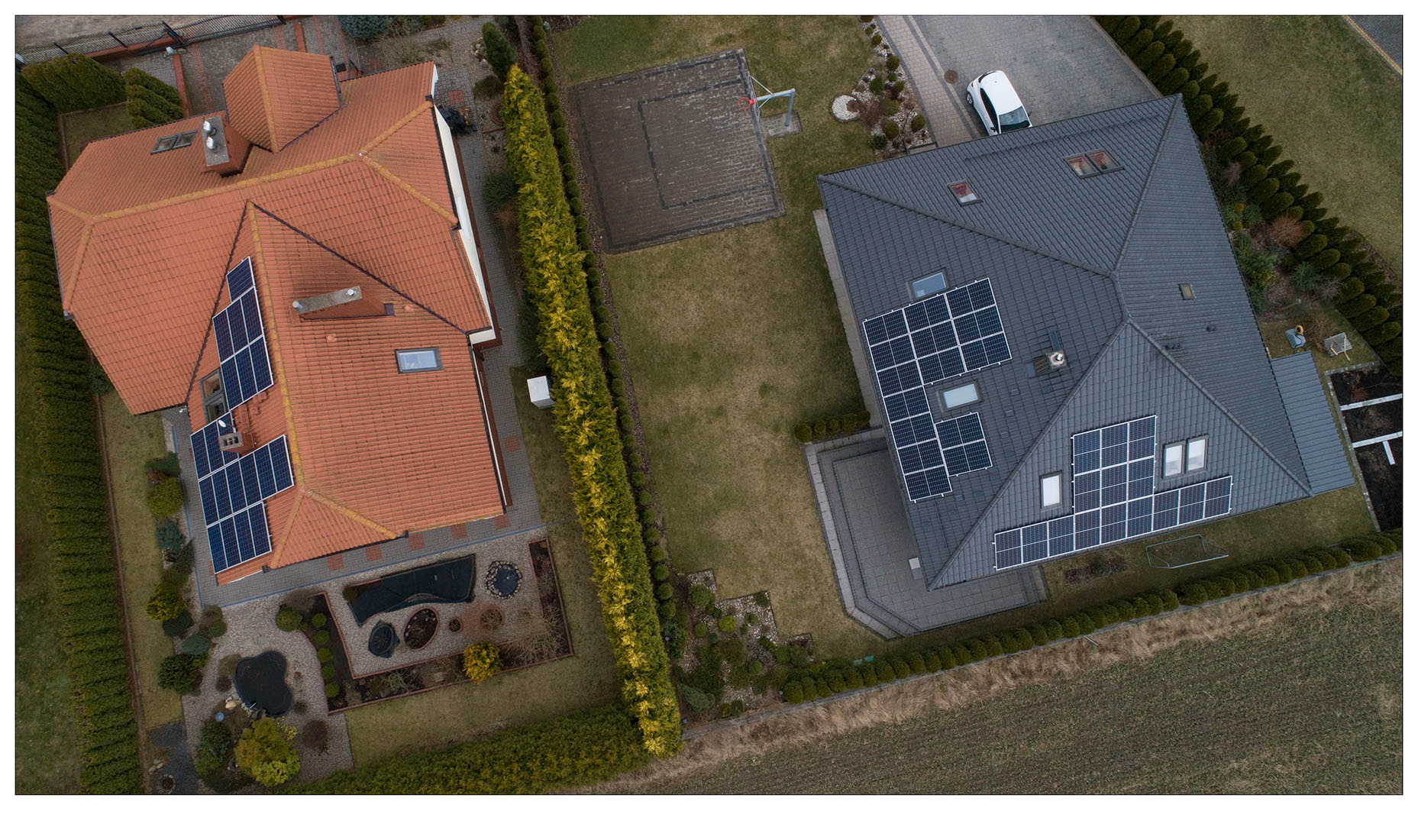 Widok z góry na dwa domy jednorodzinne z panelami słonecznymi na dachach, oddzielone żywopłotem, z widocznym boiskiem do koszykówki i samochodem.