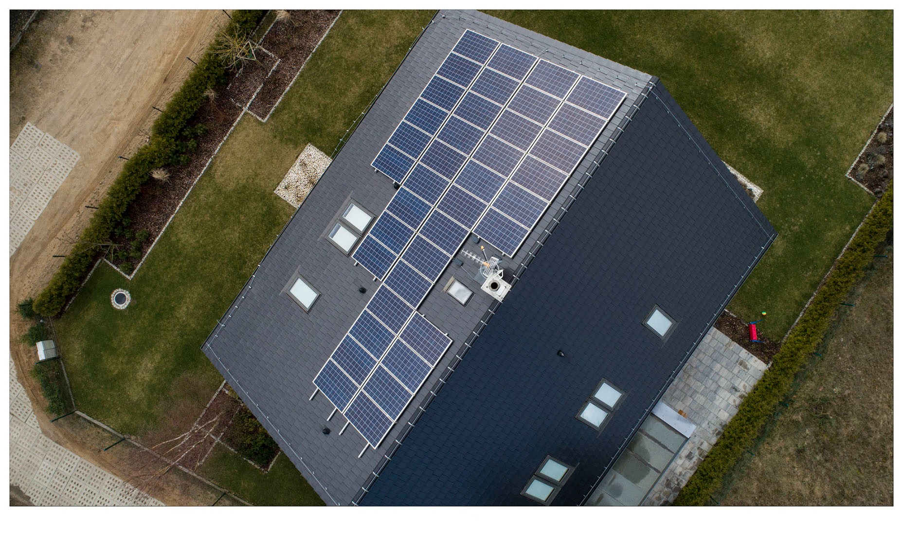 Widok z drona na nowoczesny dom z ciemnym dachem pokrytym panelami słonecznymi i kilkoma oknami dachowymi, otoczony zadbanym trawnikiem i ogrodzeniem z żywopłotu.