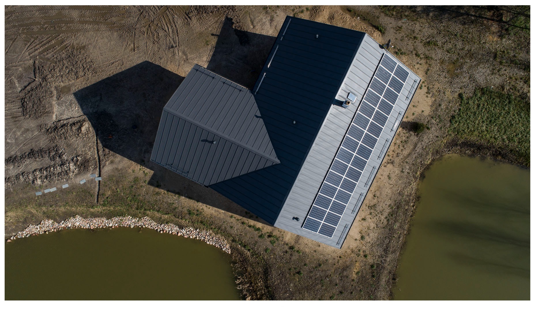 Widok z drona na dom z szarym dachem i panelami słonecznymi, obok zbiornik wodny z kamiennym brzegiem, teren częściowo zagospodarowany.