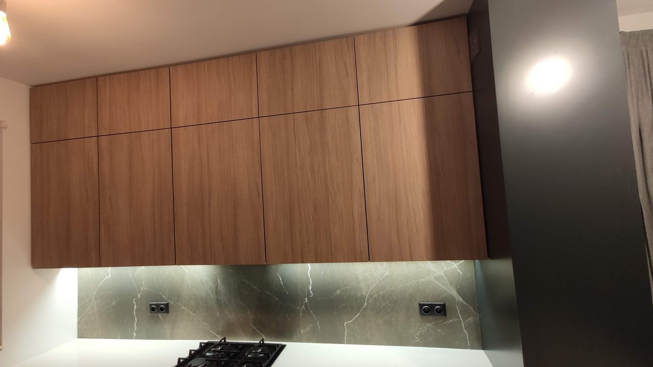 Drewniane szafki kuchenne wiszące nad blatem z marmurowym panelem i płytą gazową, oświetlenie podszafkowe, ściana w odcieniach szarości.