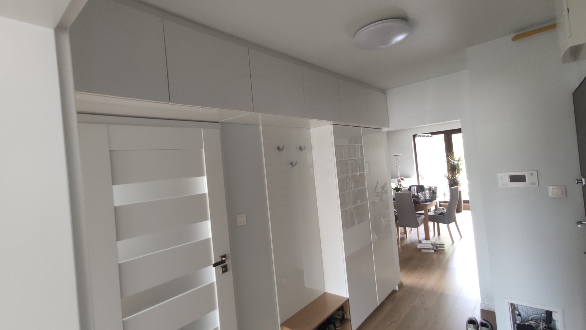 Biała szafa wnękowa z lustrzanymi drzwiami i wieszakami w przedpokoju, widoczny fragment drzwi z poziomymi przeszkleniami oraz częściowo widoczna jadalnia z oknem.