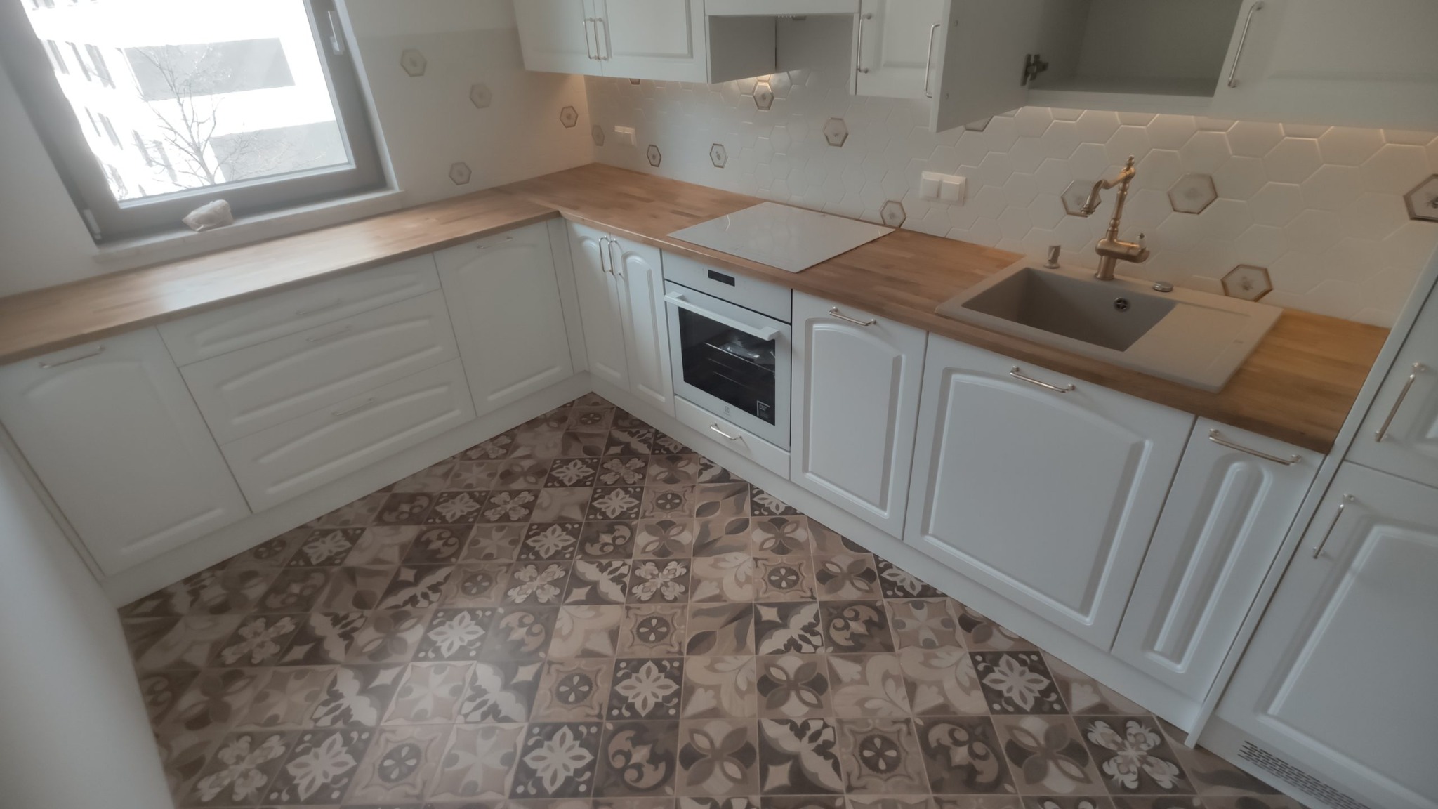 Jasna kuchnia z białymi szafkami w stylu retro, drewnianym blatem i wzorzystą podłogą. Widoczny zlewozmywak z ozdobną baterią oraz piekarnik.