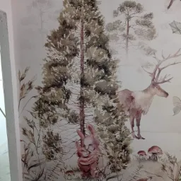 Fragment ściany z fototapetą przedstawiającą leśny krajobraz z motywami zwierzęcymi, utrzymaną w pastelowych, stonowanych barwach. Widoczne drzewa iglaste, jelenie, króliki i ptaki, tworzące spójną...