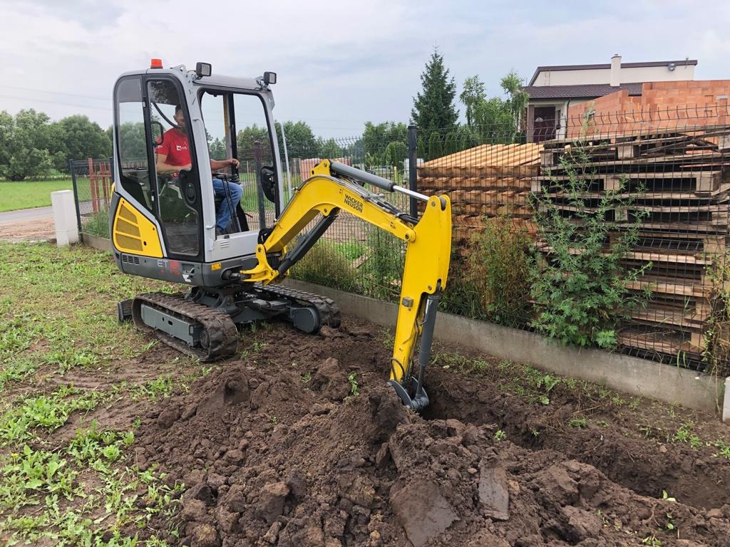 Operator w czerwonej koszulce obsługuje minikoparkę Wacker Neuson ET1, która wykonuje niewielki wykop w ziemi na trawiastym terenie, w tle widoczne palety opierające się o ogrodzenie z siatki.