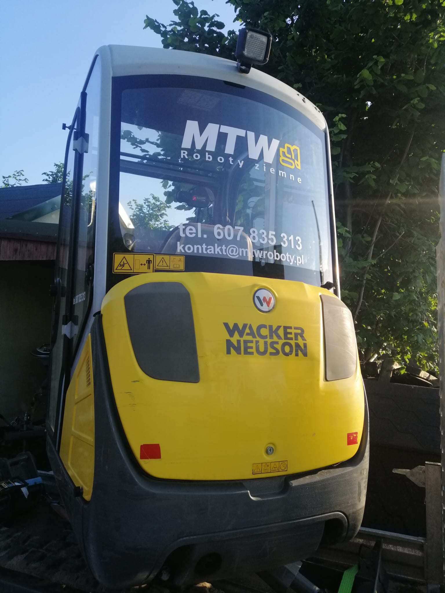 Żółto-szara minikoparka Wacker Neuson z widocznym logo MTW Roboty Ziemne i danymi kontaktowymi na szybie kabiny, stojąca na tle zieleni i fragmentu budynku.