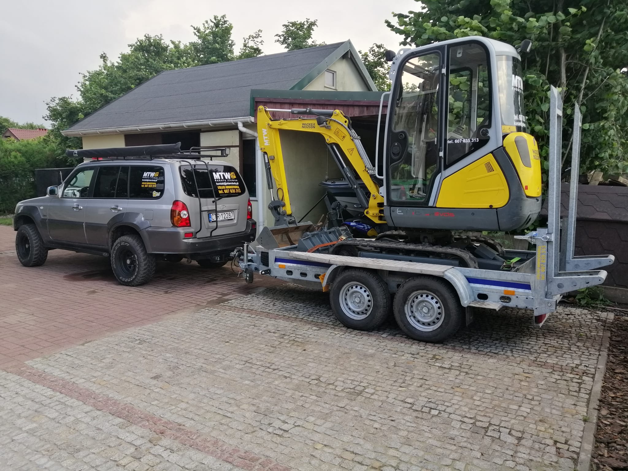 Żółto-szara mini koparka Wacker Neuson na niskopodwoziowej przyczepie, holowana przez szary samochód terenowy z naklejkami firmy budowlanej MTW na tylnej szybie, zaparkowane na kostce brukowej...