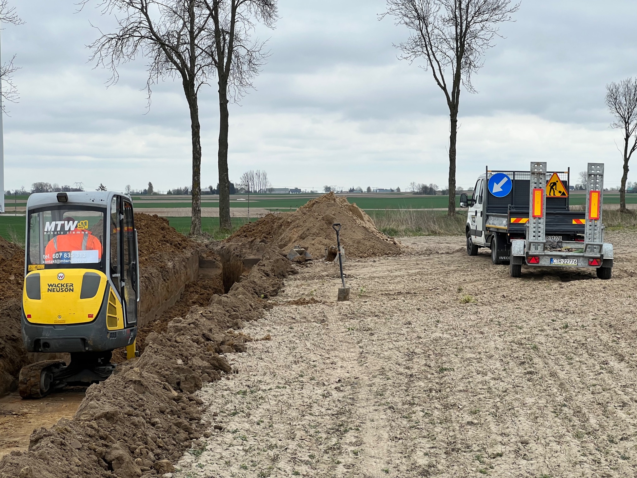 Żółto-szara mini koparka Wacker Neuson przy wykopanym wąskim rowie w polu, obok sterta ziemi i łopata, w tle biały samochód dostawczy z przyczepą i znakami ostrzegawczymi.