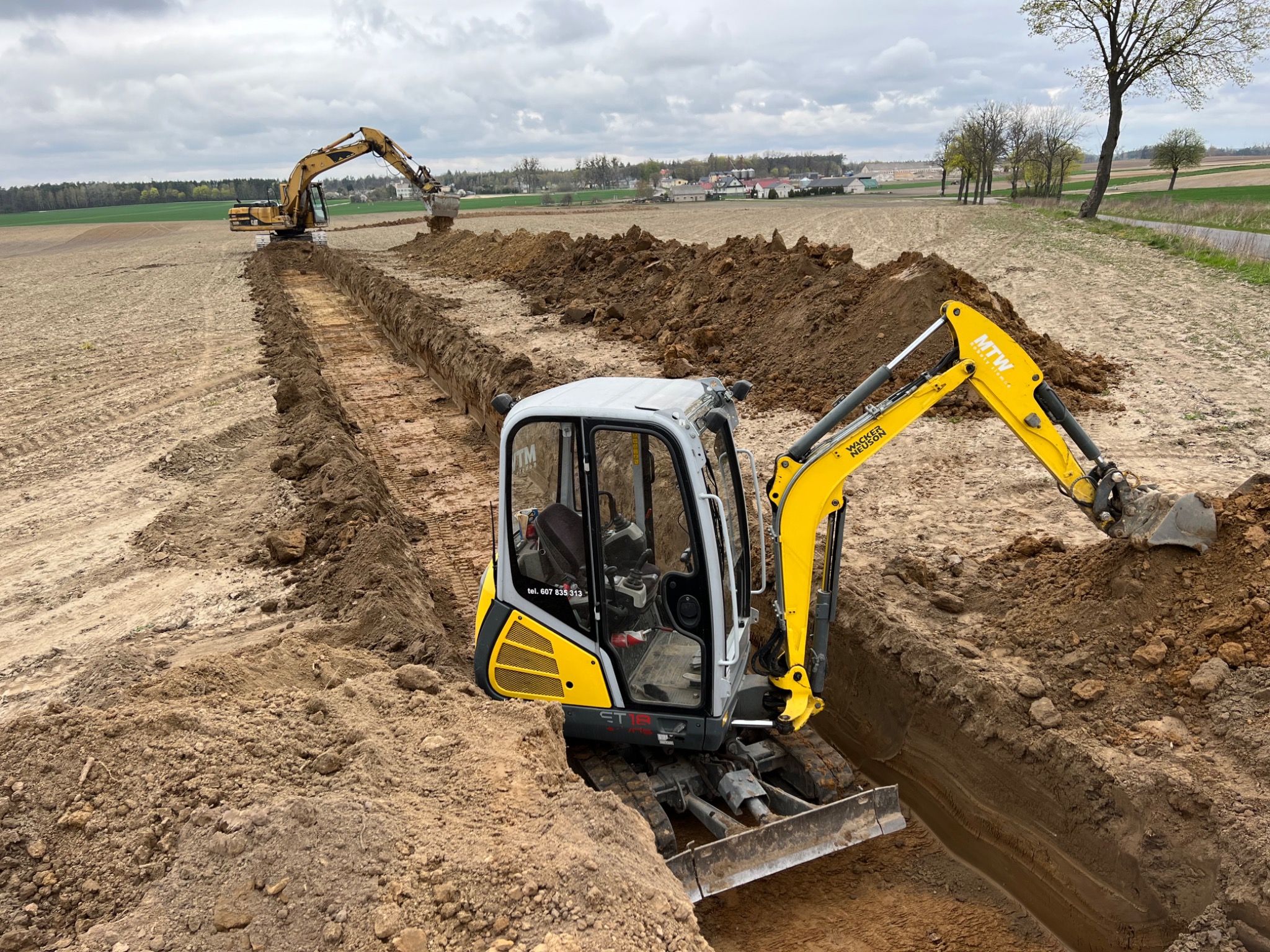 Dwa żółte baggery pracują na polu, kopiąc długi, prosty rów pod pochmurnym niebem. Mniejszy bagger znajduje się na pierwszym planie, a większy w oddali, kontynuując wykop. Ziemia wyrzucona z rowu...