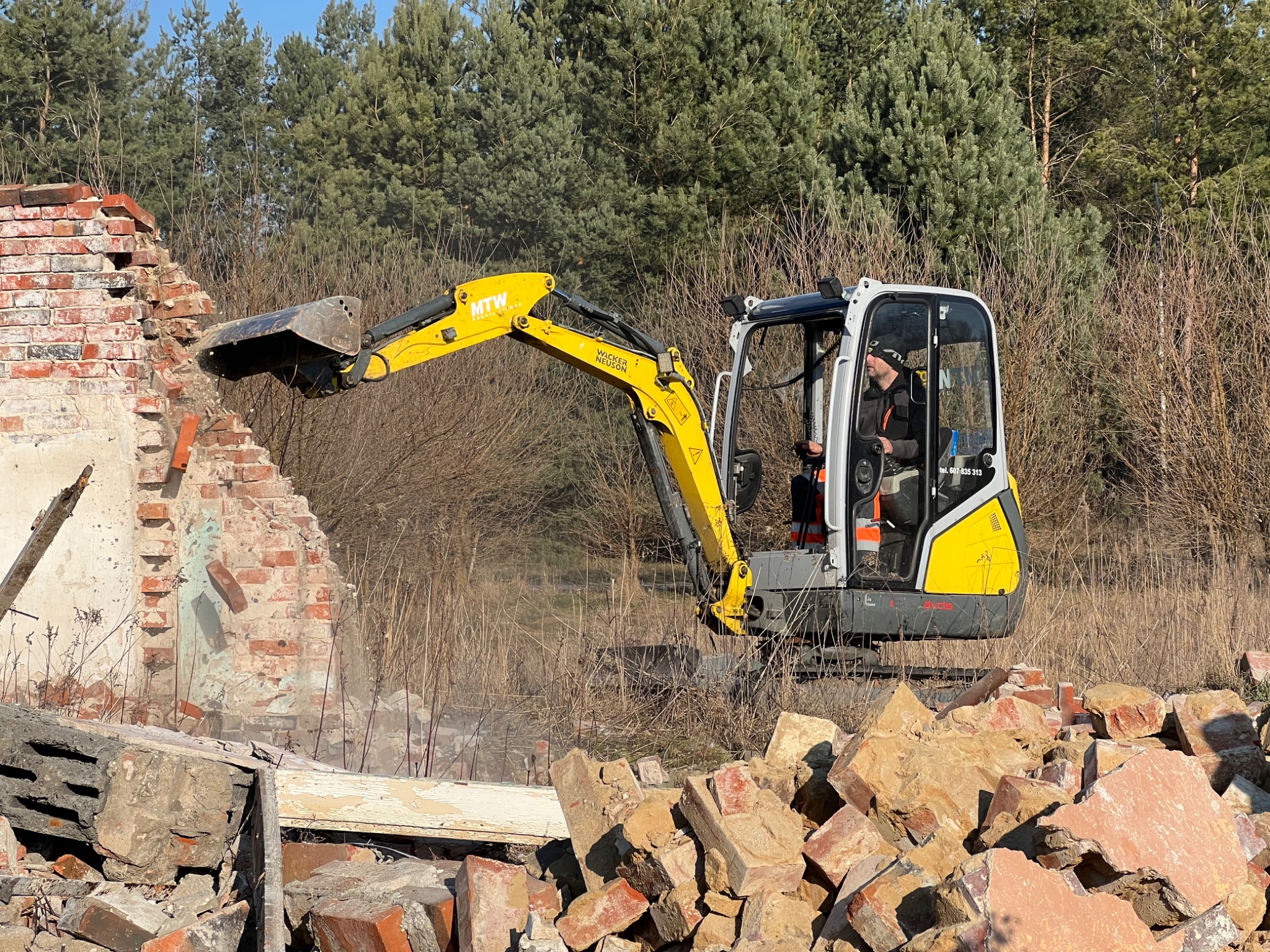 Żółta mini koparka Wacker Neuson burzy resztki ceglanego budynku na tle drzew.