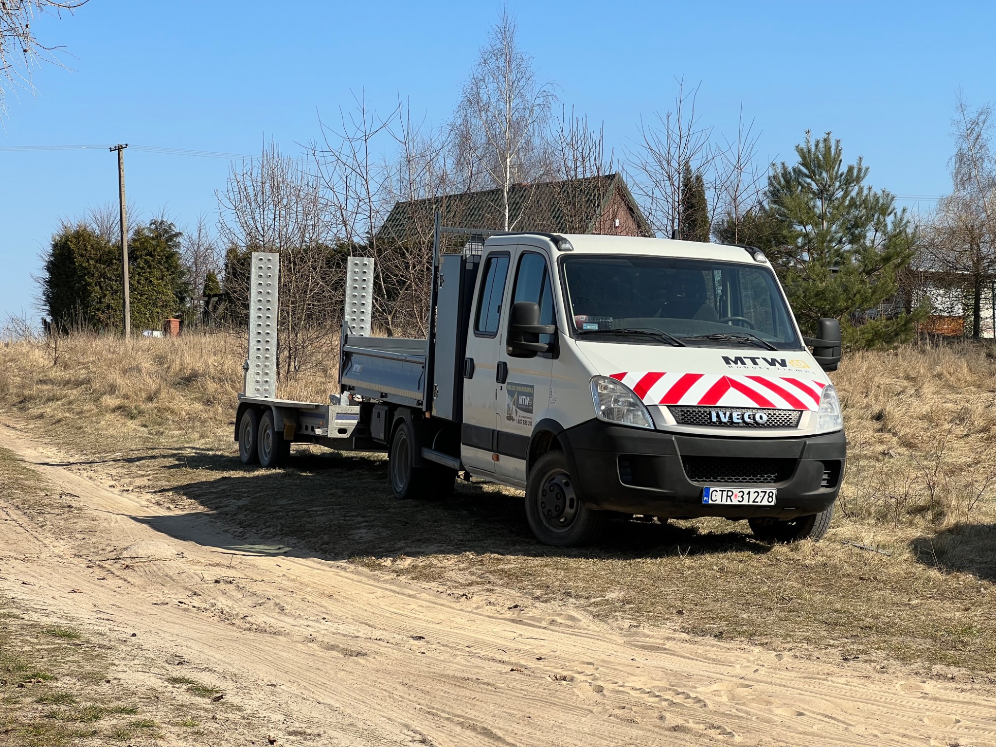 Biała ciężarówka Iveco z przyczepą na piaszczystej drodze w terenie niezabudowanym, w tle budynek z ciemnym dachem i drzewa bez liści.
