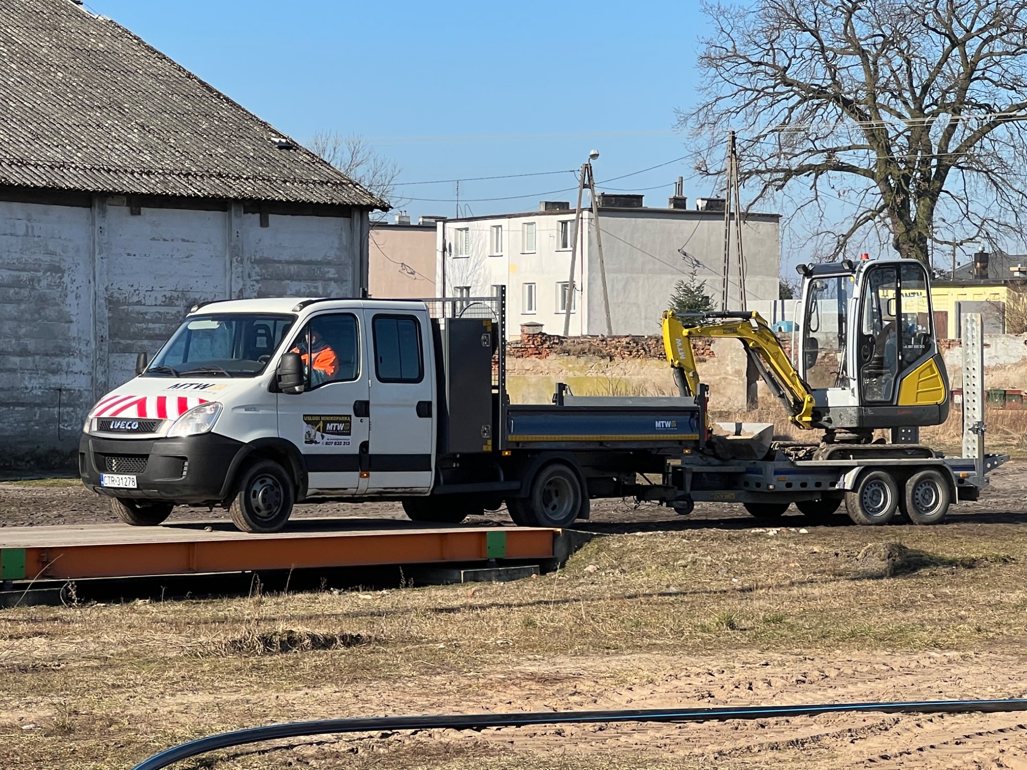 Biały samochód dostawczy Iveco z przyczepą, na której stoi żółta mini koparka, na tle zabudowań i drzew, w słoneczny dzień.