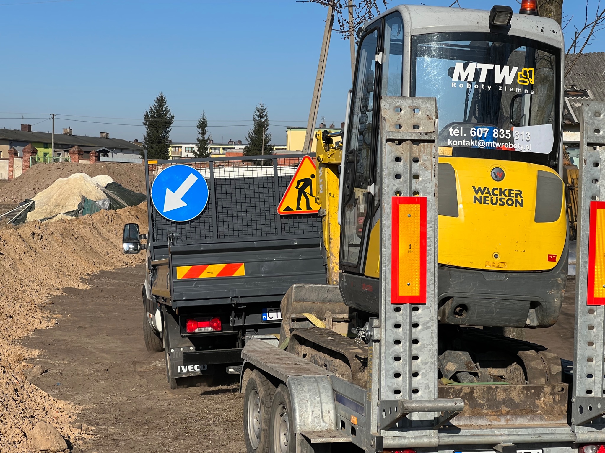 Żółta mini koparka Wacker Neuson na niskopodwoziowej przyczepie załadowana na ciężarówkę Iveco na placu budowy, widoczny znak ostrzegawczy o robotach drogowych i sterta ziemi w tle.