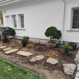 Lendar - Pracownik w trakcie sadzenia drzewek ozdobnych w ogrodzie przy białym domu. Widoczne kamienne płyty chodnikowe i narzędzia ogrodnicze.