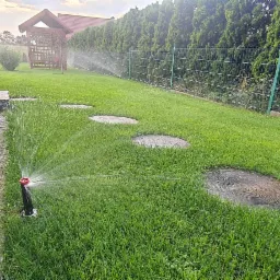 Automatyczny system nawadniania trawnika w akcji, z widocznymi okrągłymi płytami chodnikowymi i zielonym ogrodzeniem w tle.