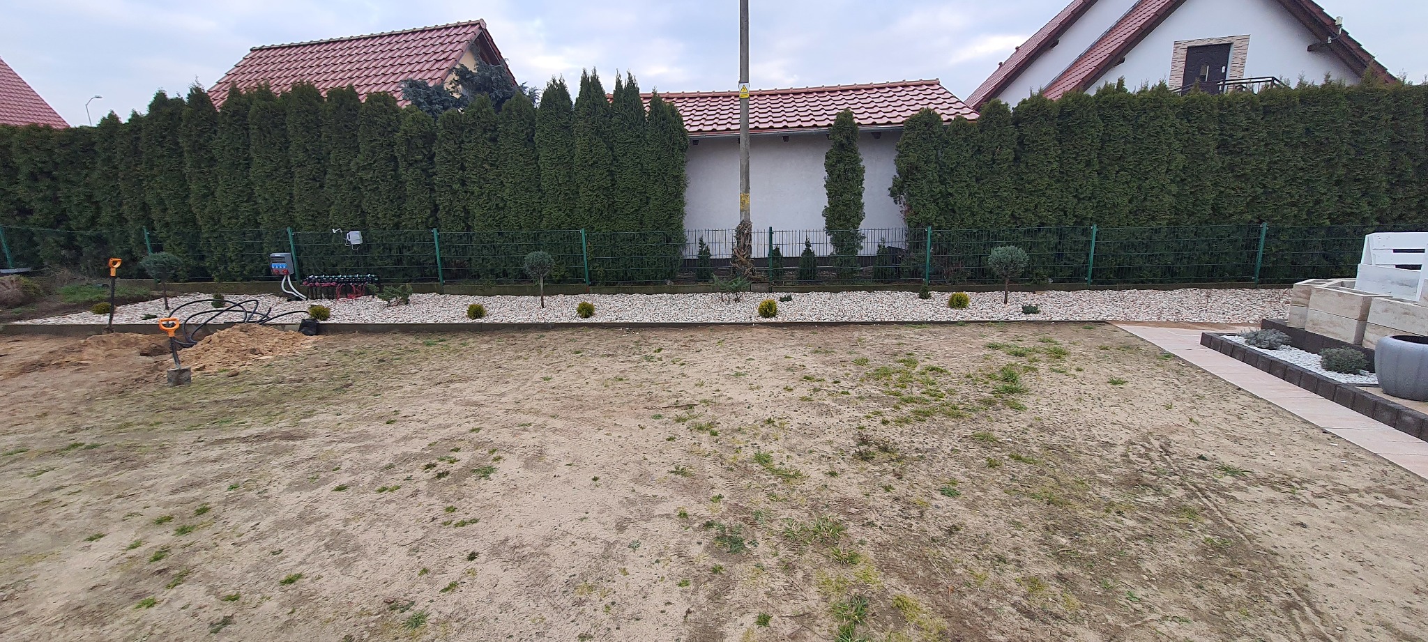 Przygotowanie terenu pod trawnik: wyrównana ziemia, system nawadniania w trakcie instalacji, widoczne szpadle i obrzeża z białego kamienia.