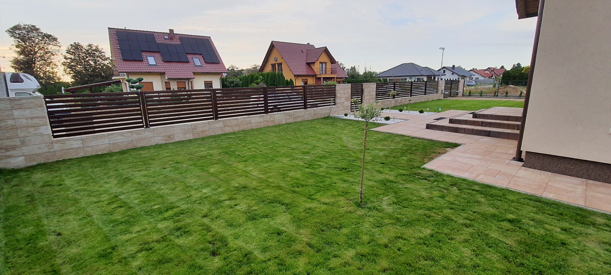 Nowo założony trawnik z młodymi drzewkami w ogrodzie z nowoczesnym ogrodzeniem panelowym i kamienną podmurówką, widok na domy jednorodzinne w tle.