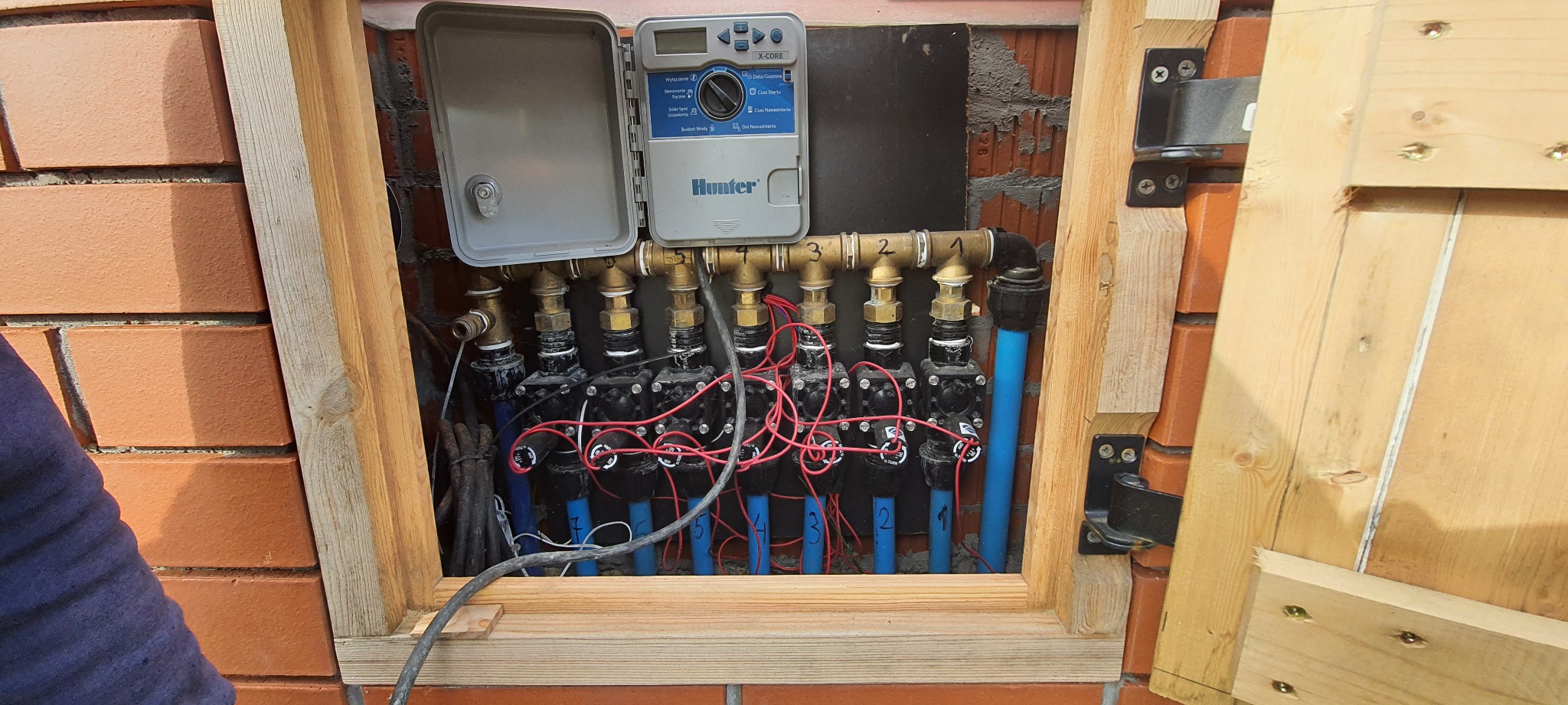 Skrzynka z kontrolerem i zaworami systemu nawadniania Hunter X-Core, widoczne rury i okablowanie, montaż w murze z obramowaniem z surowego drewna.