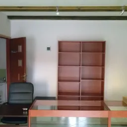 Jasne biuro po odświeżeniu: biurko z blatem z przezroczystego szkła, regał z półkami z brązowego drewna, czarne krzesło biurowe, widok na umywalkę w otwartej łazience, listwa oświetleniowa...